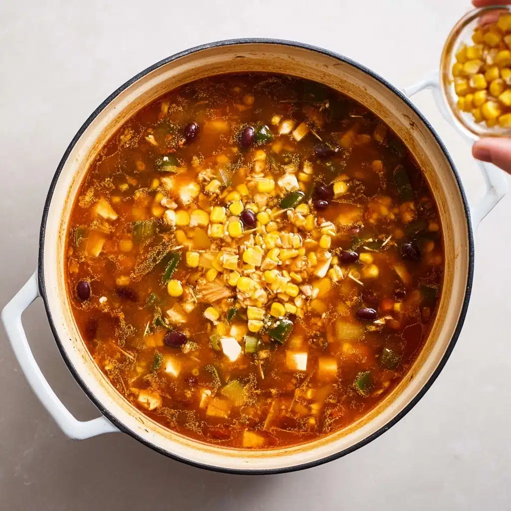 A round white Dutch oven rests on a light-colored countertop, filled with a reddish-brown broth containing visible chunks of vegetables, dark beans, and small cubes of white cheese or tofu scattered throughout. The uppermost layer shows freshly added bright yellow corn kernels tumbling in from a small glass bowl held by a hand on the right, creating motion and contrast near the surface. The soup appears hearty with a mix of green, yellow, white, and dark specks floating in the broth, all surrounded by the light interior of the pot, with no distinct separation between layers, but rather an even distribution of ingredients throughout the liquid. photo taken with an iphone
