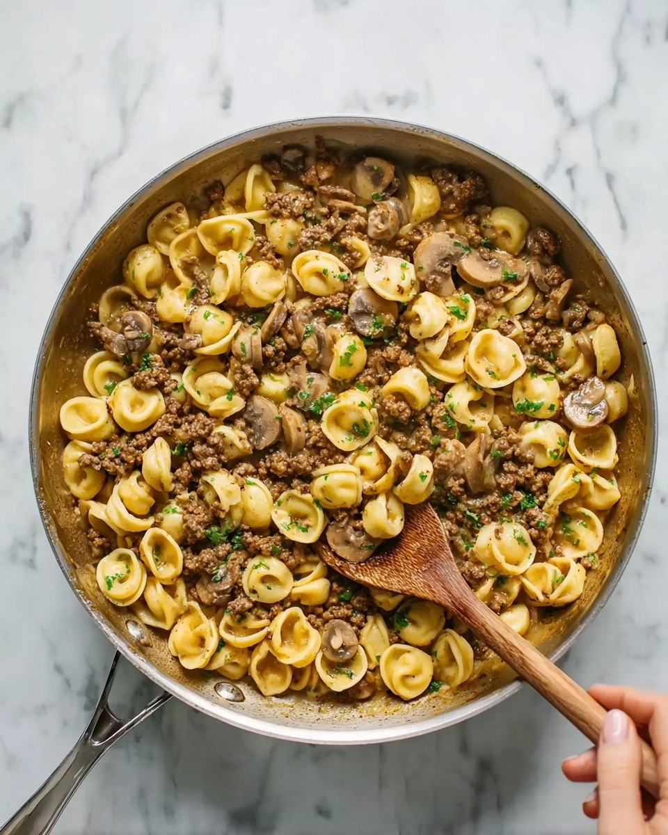 A large silver pan filled with a cooked pasta dish showing two main layers: the bottom layer is a light brown sauce mixed with small pieces of browned ground meat, and the top layer is small, round orecchiette pasta pieces that are yellow with a smooth texture. Slices of cooked mushrooms add an earthy brown tone scattered evenly throughout. Light green herb bits are sprinkled across the dish, adding a fresh color contrast. A wooden spoon is stirring the mixture from the right side, held by a woman's hand. The pan rests on a white marbled surface. photo taken with an iphone --ar 4:5 --v 7