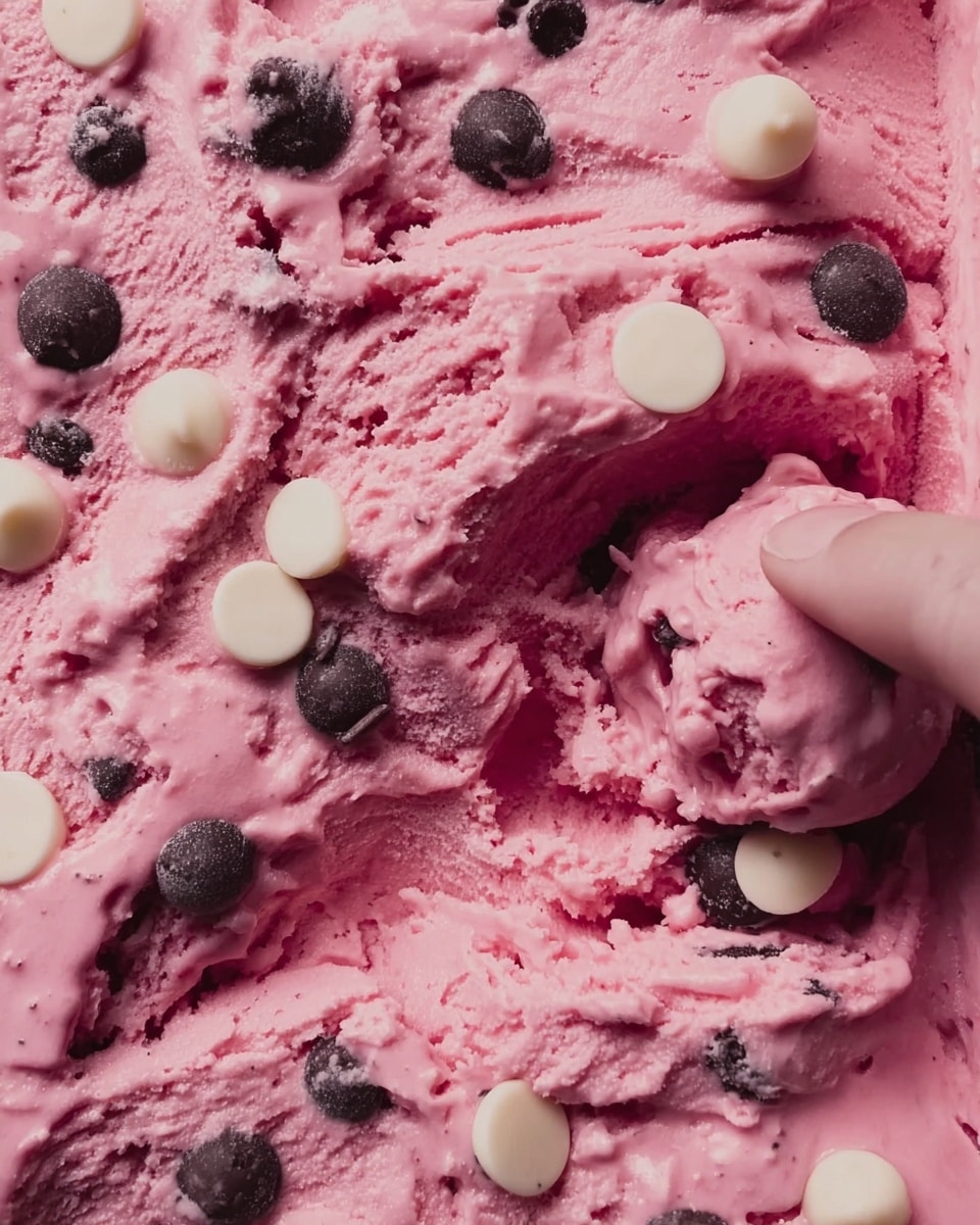The image shows a close-up of pink ice cream filled with round dark chocolate and white chocolate chips scattered throughout. The texture of the ice cream is smooth but thick, with visible scoops taken from it. A woman's hand is seen holding a scoop of the ice cream, pressing into it, causing small folds and creases. The background features a white marbled texture. photo taken with an iphone --ar 4:5 --v 7