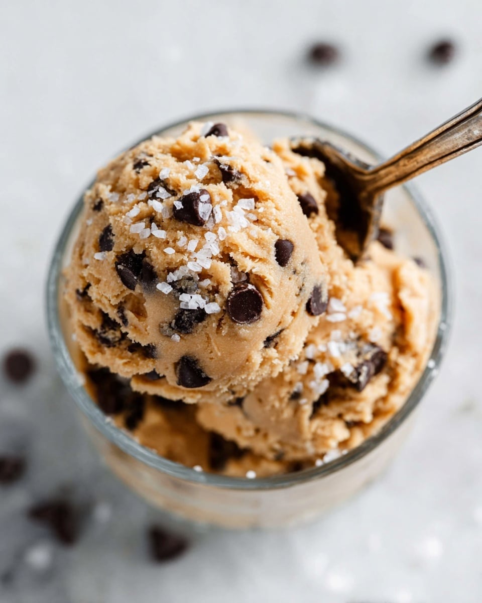 A close-up view of a small glass bowl filled with two scoops of light brown cookie dough that has many small dark brown chocolate chips mixed in. The top layer of dough is sprinkled with coarse white salt flakes, adding texture and contrast. A silver spoon is partially inserted into the dough at an angle from the upper right, resting on the top scoop. The bowl is placed on a white marbled surface with a few scattered chocolate chips blurred in the background. photo taken with an iphone --ar 4:5 --v 7