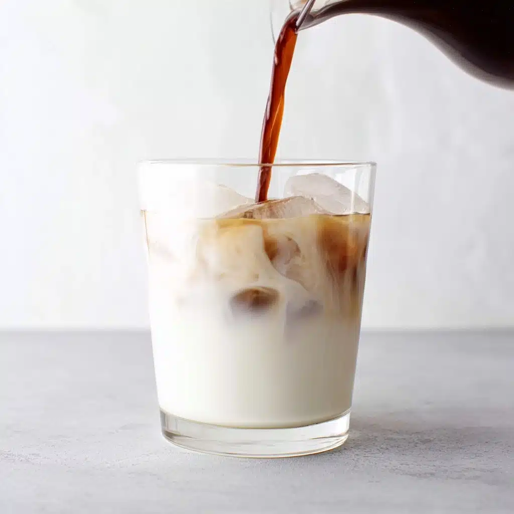 A transparent glass is filled almost to the top with a layer of cold, opaque white milk, which is packed with several clear and slightly frosty ice cubes, making the drink look extra chilled. Over the milk, a dark brown stream of coffee is being poured from a clear carafe, creating thin, swirling ribbons where the two liquids meet in the upper portion of the glass, while the rest of the drink remains solid white. The background is minimal and light, with a gray-toned surface beneath the glass and a soft, white backdrop, giving the photo a clean, fresh, and modern appearance. photo taken with an iphone