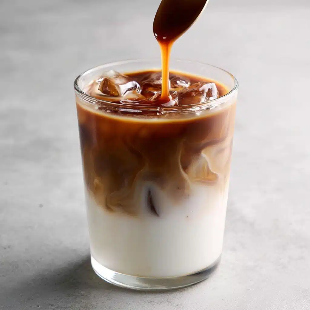 A transparent glass filled with a two-layered drink sits on a smooth grey surface, featuring a thick bottom layer of creamy white milk with visible ice cubes and a sharp transition to a rich, dark brown coffee layer on top, while a shiny stream of caramel sauce drizzles from a spoon, pooling slightly and creating a glossy ripple effect on the surface; the overall texture is cool and enticing with defined layers and a reflective, frosty appearance. photo taken with an iphone