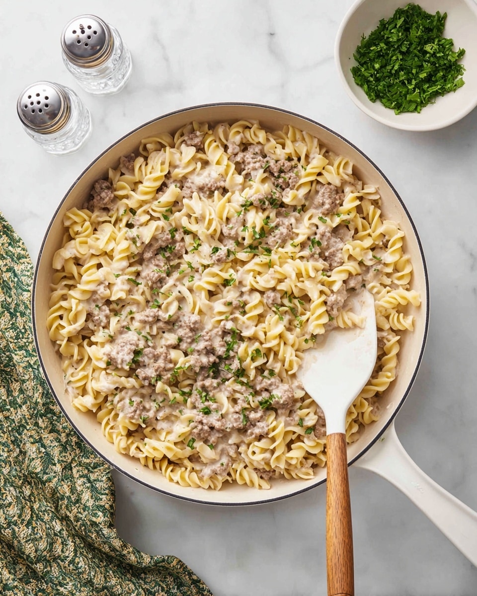 A large white pan filled with creamy pasta showing three layers: a base layer of twisted egg noodles in a pale yellow shade, a middle layer of browned ground meat scattered unevenly, and a top layer of creamy sauce mixed with the noodles and meat, creating a smooth, light beige texture. Small green parsley bits are sprinkled over the pasta adding fresh specks of color. A white spatula with a wooden handle rests inside the pan, partially lifting some pasta from the right side. The pan sits on a white marble surface next to a small white bowl filled with chopped green herbs and two clear salt and pepper shakers. A patterned green and white cloth lies at the bottom left corner of the image. Photo taken with an iphone --ar 4:5 --v 7
