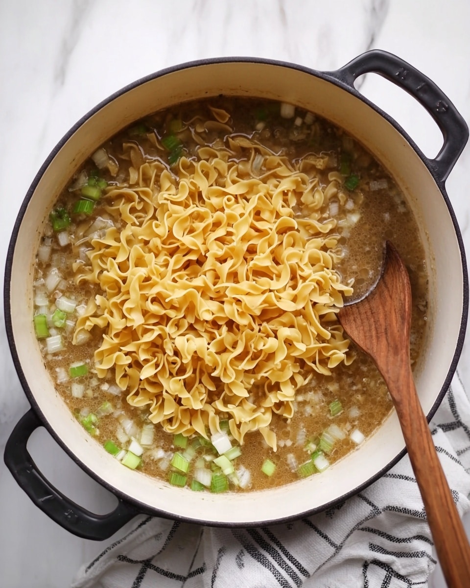 A large white pot with black handles sits on a white marbled surface, filled with light brown broth containing diced green celery and white onions. On top, there is a thick layer of uncooked yellow curly egg noodles, partially covering the broth. A wooden spoon is inside the pot, resting near the noodles, showing light wood grain texture. A white cloth with black stripes lies next to the pot. Photo taken with an iphone --ar 4:5 --v 7