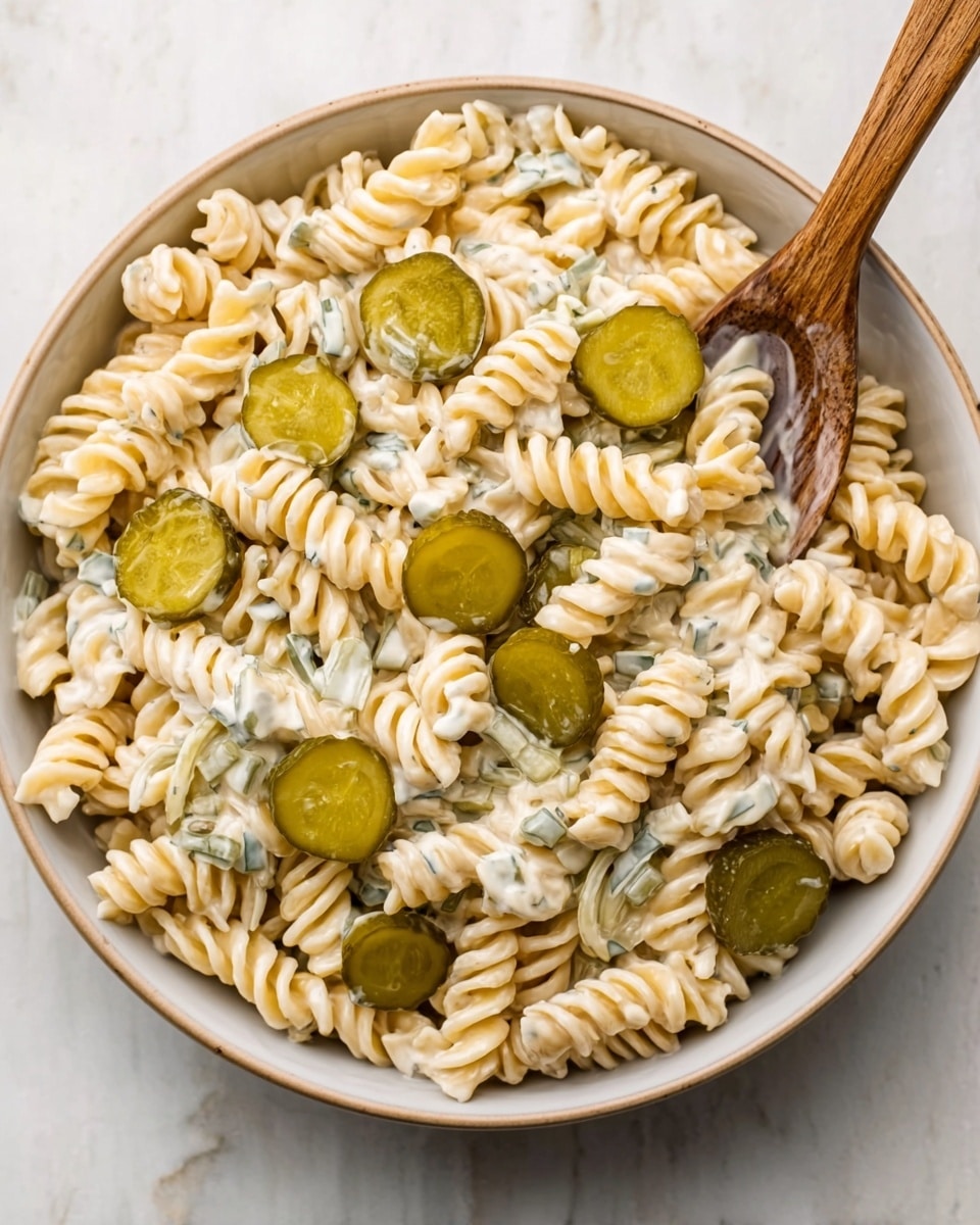 A white bowl sits on a white marbled surface, filled with a pasta salad made of twisted pasta pieces that are creamy off-white in color. Mixed throughout are small green pickle slices, each one round and evenly spread on top of the pasta. A small wooden spoon is placed inside the bowl, touching some pasta on one side. The texture of the pasta looks soft and creamy with bits of dressing visible, and the pickle slices add a pop of smooth, glossy green. photo taken with an iphone --ar 4:5 --v 7