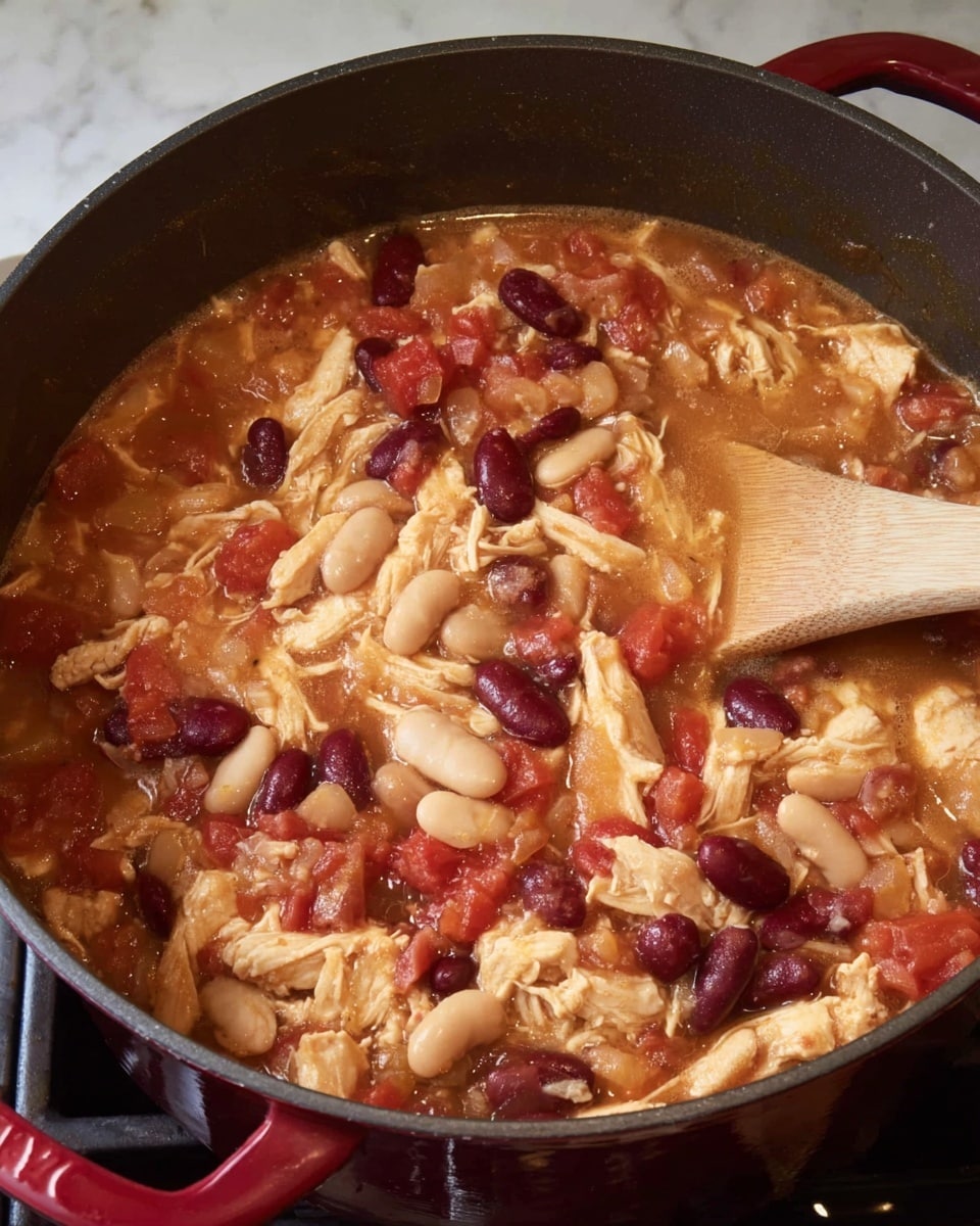 A close-up view of a thick stew in a deep black cooking pot, filled with shredded light brown chicken pieces, white beans, deep red kidney beans, and chunks of bright red tomatoes mixed in a reddish broth. The stew shows a textured mix of soft beans and tender meat pieces, with a wooden spoon partially visible stirring on the right side of the pot. The pot sits on a dark stove with a white marbled backdrop around it. Photo taken with an iphone --ar 4:5 --v 7