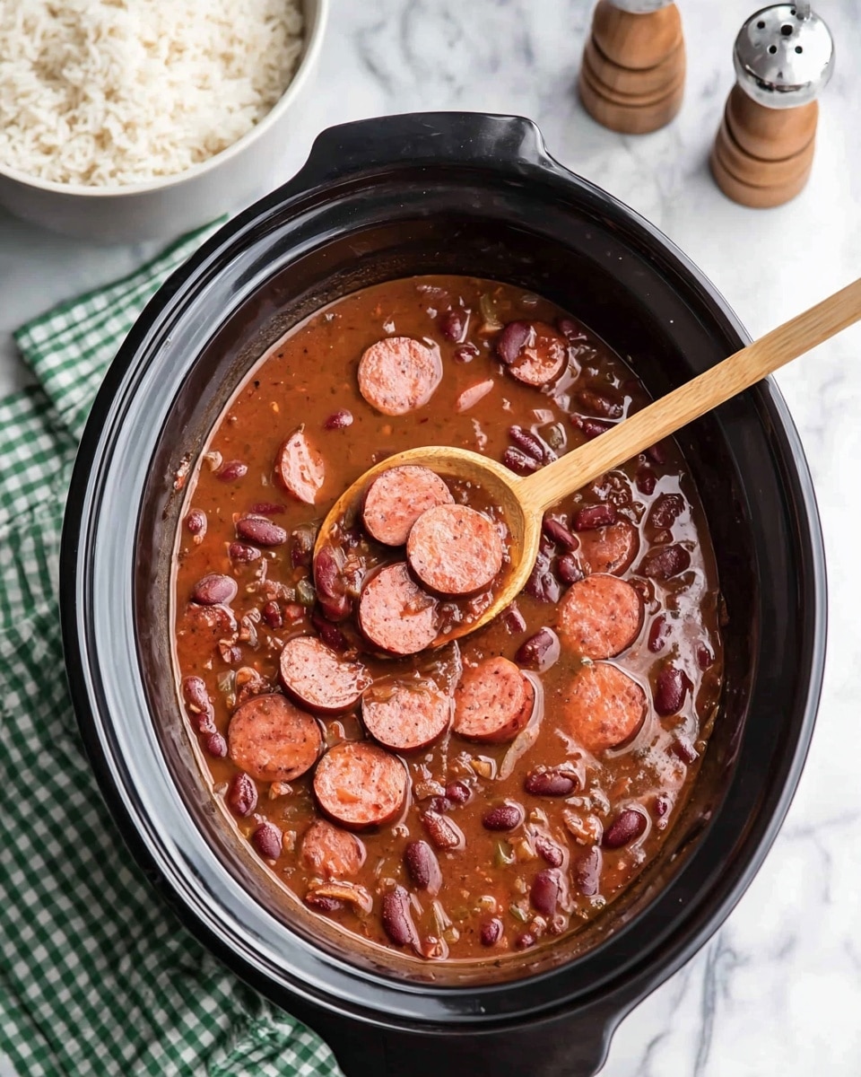 A black slow cooker filled with thick reddish-brown stew containing sliced sausage and red beans, with a wooden spoon resting inside holding several sausage slices. The slow cooker is placed on a white marbled surface with a green and white checkered cloth beside it. In the top left corner, there is a white bowl filled with cooked rice, and in the top right corner, salt and pepper shakers are visible. photo taken with an iphone --ar 4:5 --v 7