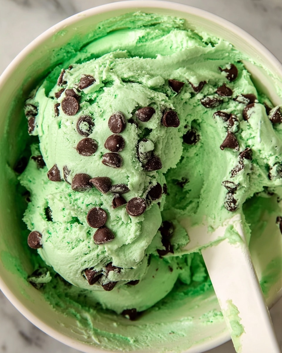 A close-up image of bright green mint chocolate chip ice cream dough with smooth, creamy texture filled with many dark brown chocolate chips scattered throughout. The ice cream dough is in a white bowl, and a white spatula covered with the green dough is resting on the right side inside the bowl. The background shows a white marbled surface. photo taken with an iphone --ar 4:5 --v 7
