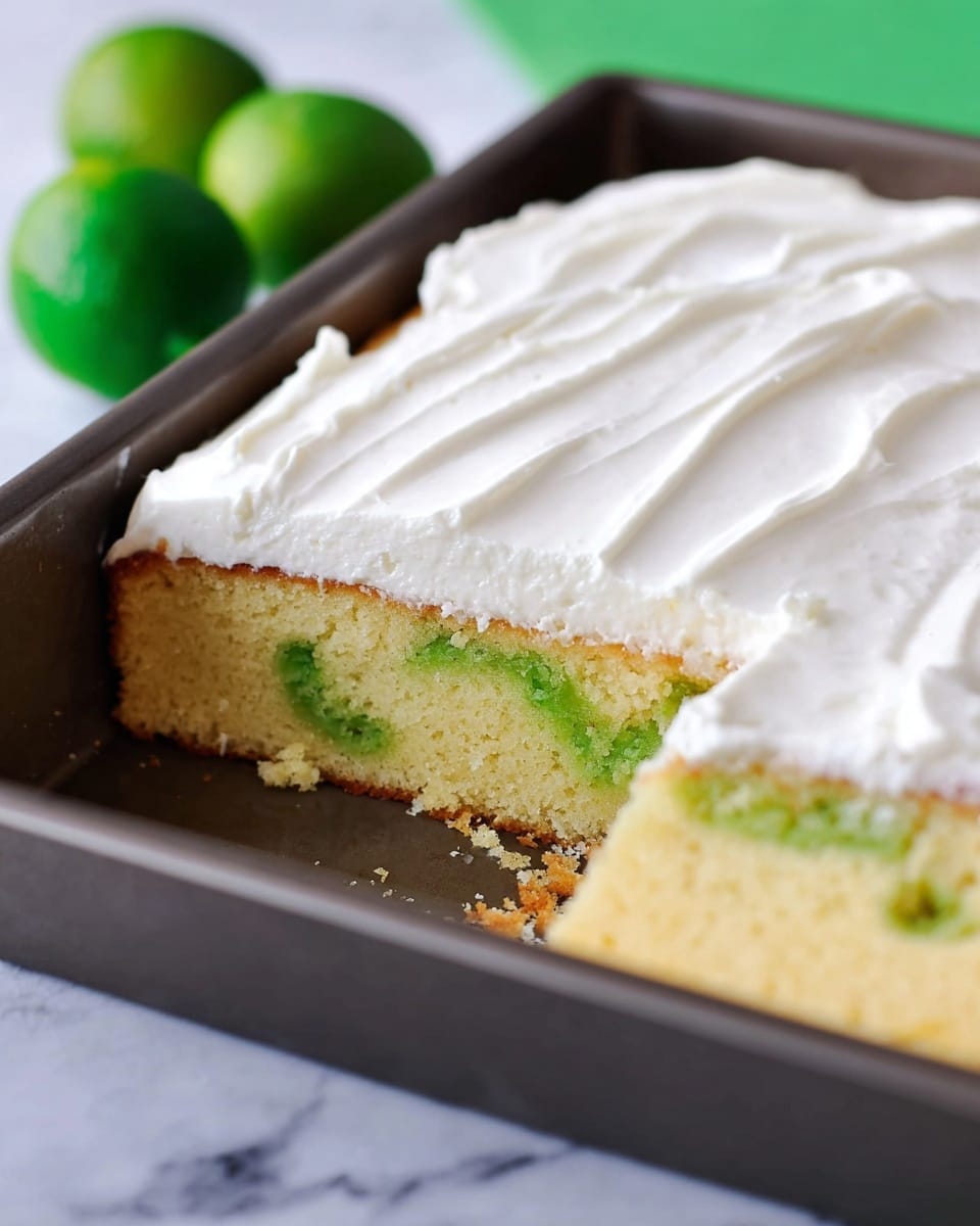 A rectangular cake in a dark baking tray with a white marbled surface underneath has one piece removed, showing two layers: the bottom layer is a light yellow sponge with green swirls inside, and the top layer is a smooth thick white cream evenly spread over the cake. The cream layer has a soft, fluffy texture and smooth edges. Two green limes are blurry in the background. Photo taken with an iphone --ar 4:5 --v 7