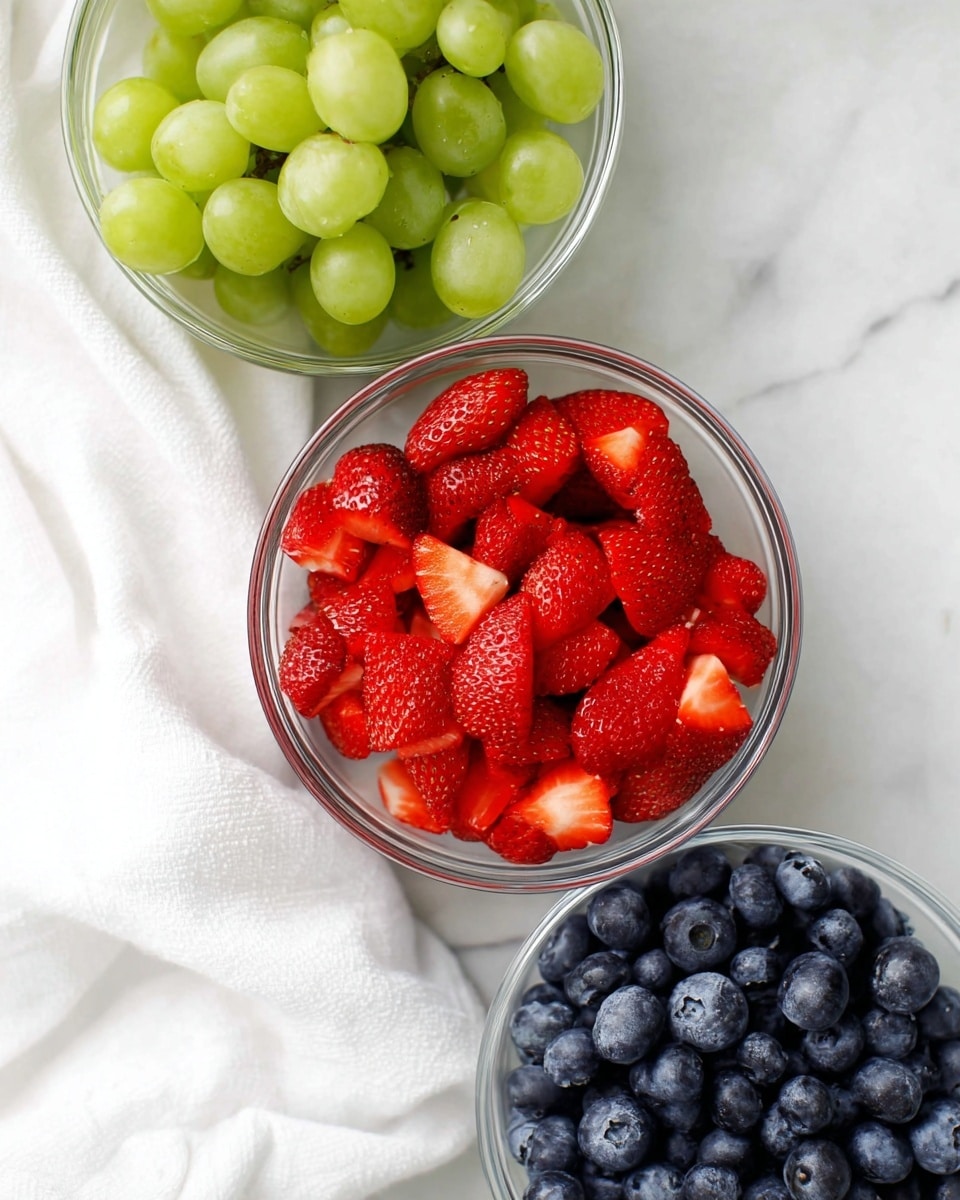 Three clear glass bowls on a white marbled surface with a soft white cloth on the left. The top bowl is filled with shiny green grapes, the middle bowl holds bright red strawberry halves with smooth, juicy texture, and the bottom bowl is full of plump, deep blue blueberries with a slightly frosted look. The bowls are arranged so that the middle bowl is centered in the image, with the other two slightly cut off at the edges. photo taken with an iphone --ar 4:5 --v 7