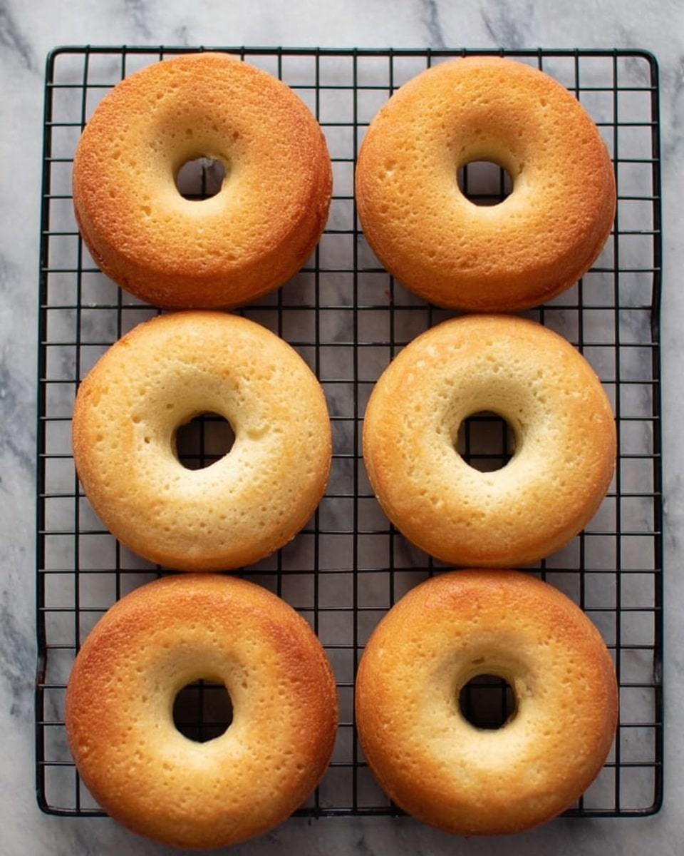 Six plain baked donuts arranged in two rows of three on a black cooling rack, placed over a white marbled surface. Each donut has a golden-brown top with a smooth texture and a slightly lighter, almost creamy-colored bottom. The donuts have round shapes with even holes in the center, showing a soft, light interior with some small air pockets. The lighting highlights the warm tones and gentle crust on each donut. Photo taken with an iphone --ar 4:5 --v 7