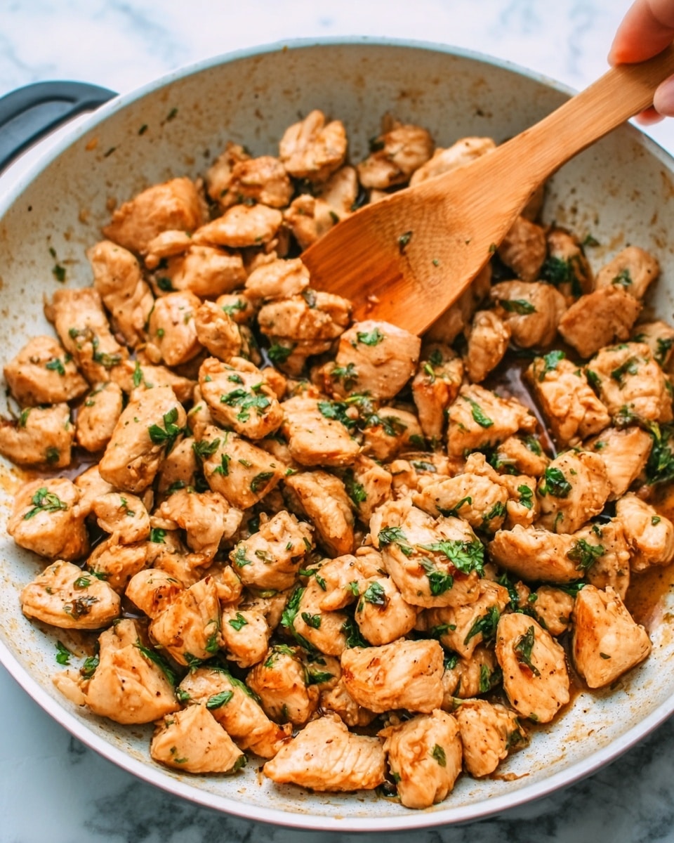 The image shows a white pan filled with many small pieces of cooked chicken that are light brown and slightly shiny, with some green herbs sprinkled on top. A woman’s hand is holding a wooden spoon stirring the chicken. The pan is placed on a white marbled surface. The chicken pieces have a soft texture and appear tender and juicy. photo taken with an iphone --ar 4:5 --v 7
