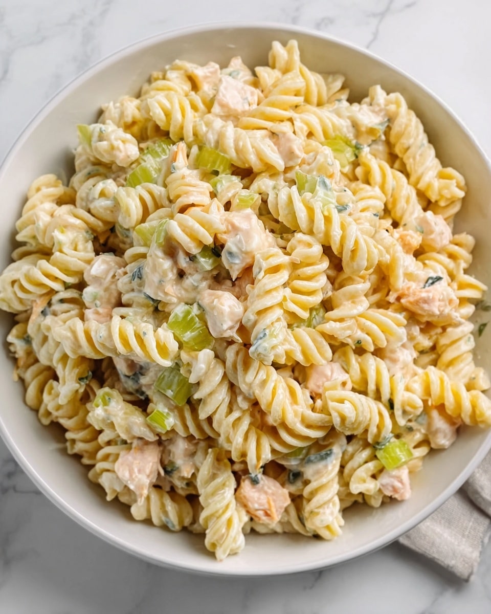 A white bowl filled with a creamy pasta salad made of spiral pasta. The pasta is pale yellow mixed with small pieces of light pink cooked chicken and bits of green celery. The salad looks creamy and soft, with a slightly shiny texture from the dressing. The bowl is placed on a white marbled surface. photo taken with an iphone --ar 4:5 --v 7