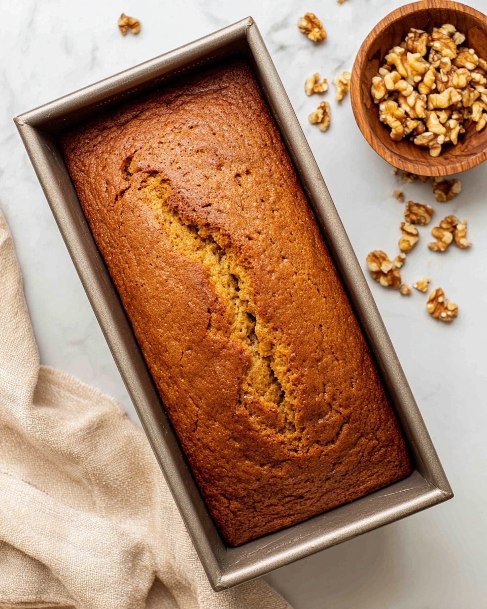 A loaf of brown cake with a cracked top sits in a metal rectangular baking pan, showing a slightly rough texture and a golden-brown color. To the upper right of the pan, a small wooden bowl filled with chopped walnuts is placed on a white marbled surface with some walnut pieces scattered around it. A light beige cloth is seen to the lower left of the pan, adding softness to the scene. The overall look is warm and inviting. photo taken with an iphone --ar 4:5 --v 7