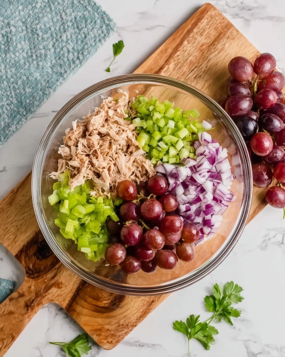 A clear glass bowl sits on a wooden cutting board, both on a white marbled surface. Inside the bowl are four separate layers of ingredients: at the top left, pale beige shredded cooked meat, next to it on the top right, small bright green chopped celery pieces, on the bottom right finely diced purple-red onion, and at the bottom left dark red halved grapes. Outside the bowl are a small bunch of whole grapes and a few green parsley leaves scattered on the board and marble surface. Photo taken with an iphone --ar 4:5 --v 7