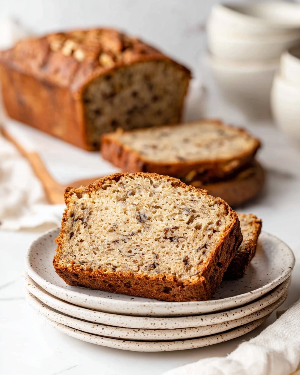 Two thick slices of light brown nut bread with visible walnut pieces and small dark specks are placed on a stack of two white plates with speckled rims. Behind the plates, a larger loaf of the same bread, partially sliced, rests on a white marbled surface, with a soft white cloth and stacked white bowls blurred in the background. The crust of the bread is darker and textured, showing a baked golden-brown color. The photo taken with an iphone --ar 4:5 --v 7