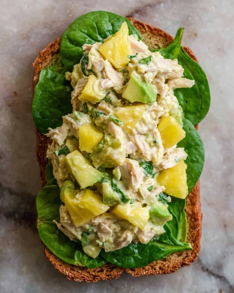 The image shows a single open sandwich on a slice of toasted brown bread. The bread is topped with fresh green spinach leaves spread evenly across the surface. On top of the spinach, there is a thick layer of a chunky, creamy mixture made of shredded light-colored tuna, light green avocado pieces, and small yellow chunks, likely mango or pineapple. The textures of the mixture are soft and slightly chunky, with bits of green herbs mixed in. The sandwich sits on a white marbled surface. photo taken with an iphone --ar 4:5 --v 7
