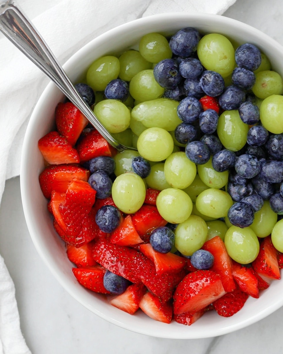 The image shows a white bowl filled with a fresh fruit mix consisting of three main layers: the first layer contains light green grapes that are smooth and shiny, scattered evenly throughout the bowl; the second layer is made up of deep blue blueberries with a slightly matte texture, filling the spaces between the grapes; the third layer has bright red strawberries cut into halves and quarters, showing their juicy interior and seeds on the surface. A silver spoon rests inside the bowl among the fruits, reflecting some of the colors. The whole bowl sits on a white marbled surface next to a white cloth. Photo taken with an iphone --ar 4:5 --v 7