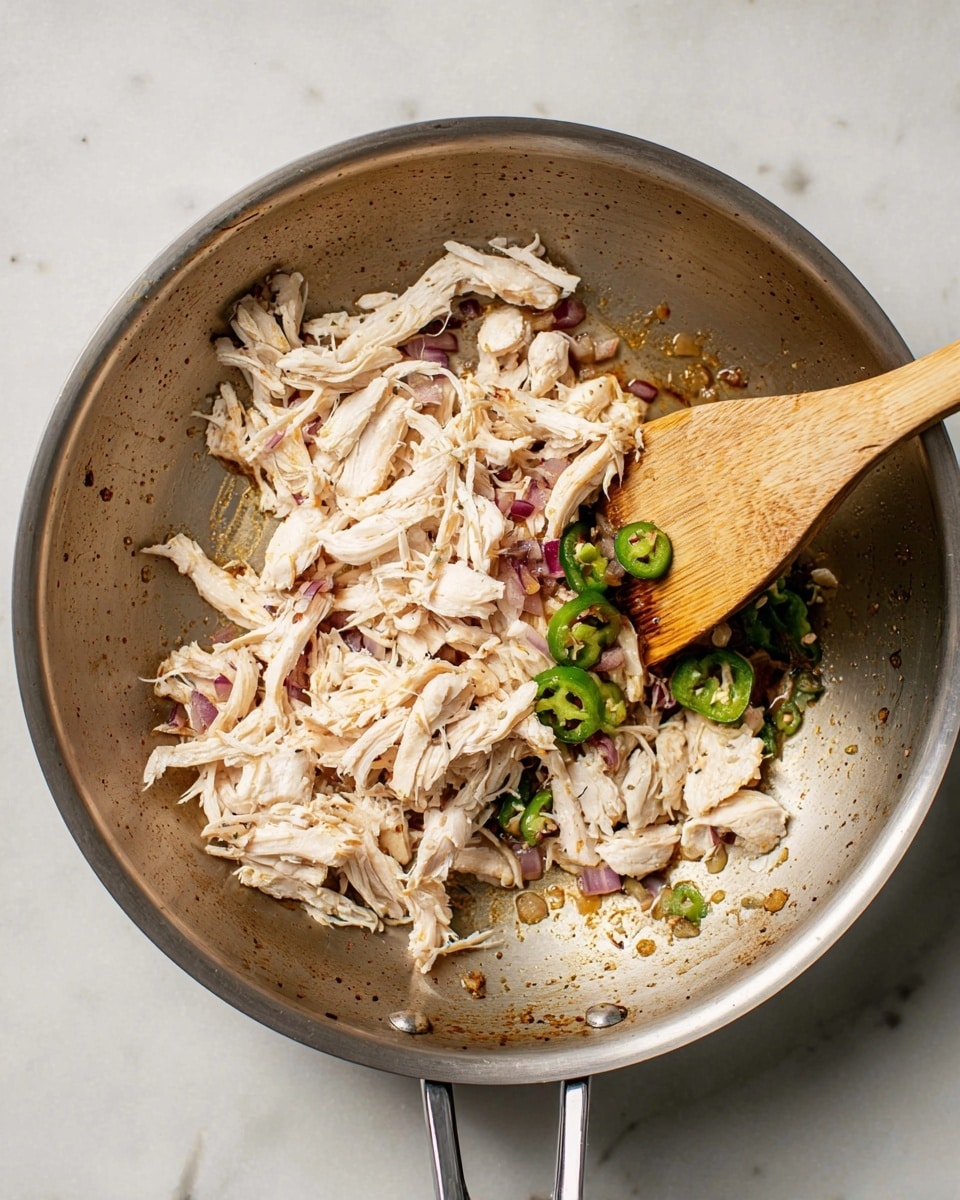 A metal pan with shredded white chicken piled mostly on the left side mixing with small pieces of diced purple onions and sliced green jalapeños near the top right edge, some browning and oil stains scattered around the pan's surface. A wooden spatula rests inside the pan, positioned diagonally from the bottom right, partially under the shredded chicken, against a white marbled textured surface. photo taken with an iphone --ar 4:5 --v 7