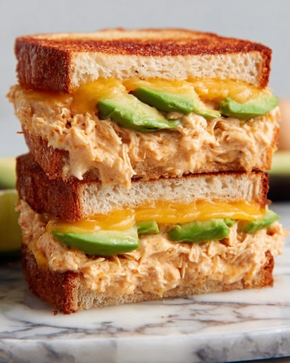 The image shows a close-up of a sandwich cut in half, stacked with three layers. The bottom layer is a piece of toasted golden-brown bread, on top of which sits a layer of light orange shredded chicken mixed with creamy sauce. Above that are slices of fresh green avocado. The middle layer is a melted orange-yellow cheese slice, slightly gooey and soft. Above the cheese is another layer of avocado slices, followed by another layer of the same shredded chicken mix. The sandwich is topped with a final piece of toasted golden-brown bread. The sandwich sits on a white marbled surface that adds brightness to the image. Photo taken with an iphone --ar 4:5 --v 7