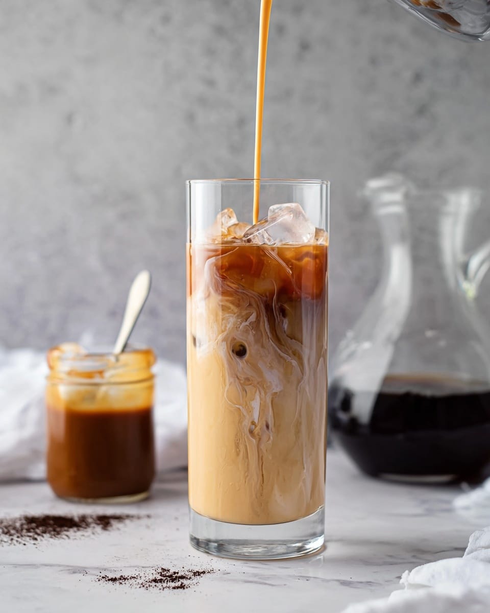 A tall clear glass filled with ice and creamy light brown iced coffee, showing layers of milk and coffee swirling together as a thin stream of darker coffee is poured into the top. The glass sits on a white marbled surface with some dark coffee grounds scattered nearby. Behind the glass to the left, there is a small glass jar with a spoon inside containing thick caramel sauce, and to the right, a clear glass coffee decanter with dark coffee inside. The background is a soft grey texture. Photo taken with an iphone --ar 4:5 --v 7