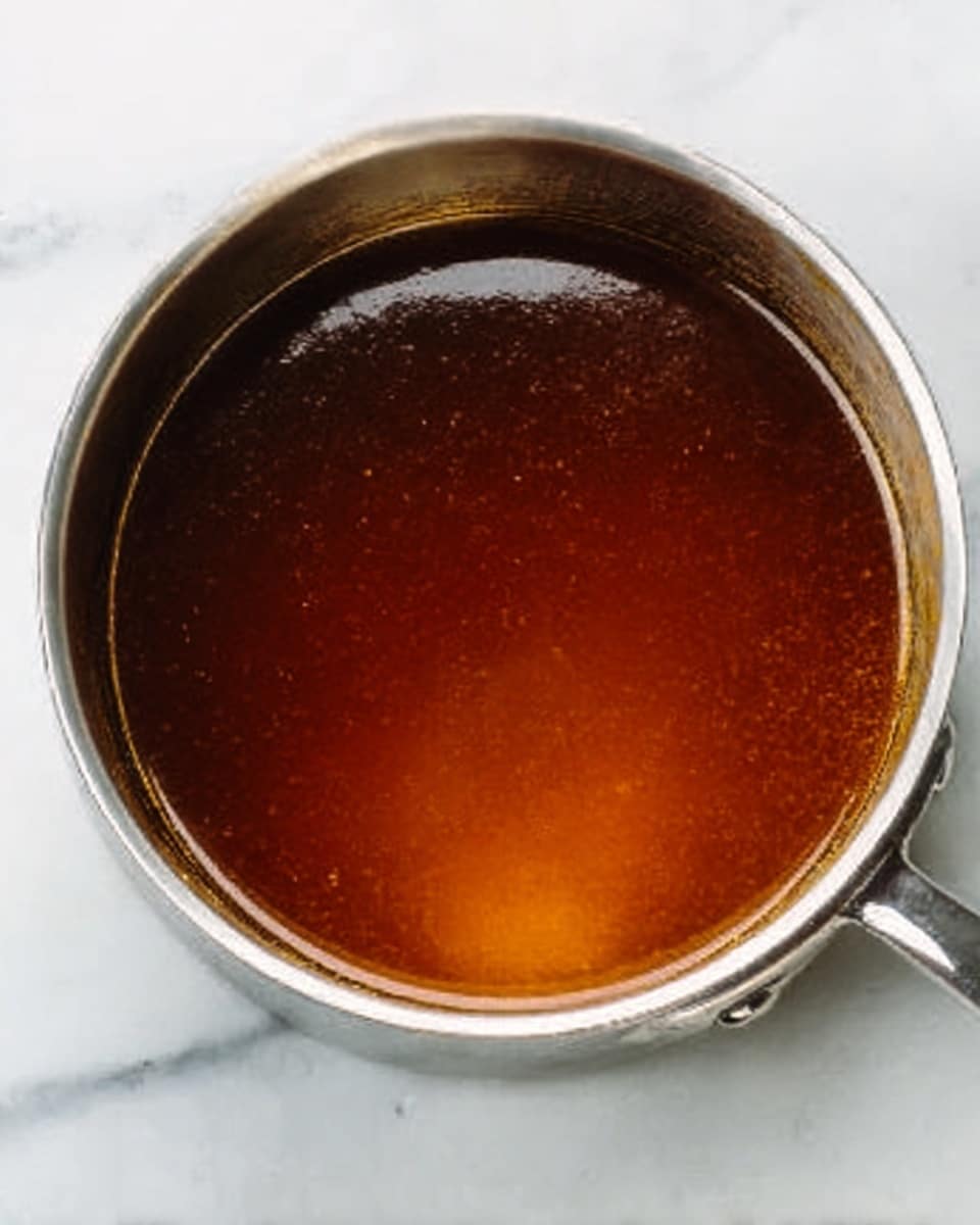 A close-up image of a round metal saucepan filled with a smooth, dark amber liquid. The liquid looks shiny and has a thick texture. The saucepan is placed on a white marbled surface, highlighting the contrast between the shiny dark liquid and the light background. The edges of the saucepan are clean, and there is a small spout visible on the side of the pan. photo taken with an iphone --ar 4:5 --v 7