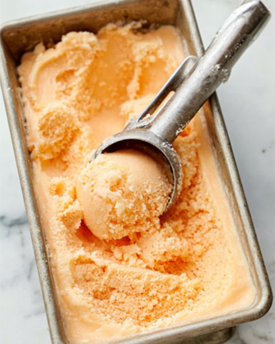 The image shows a close-up of a metal container filled with pale orange sherbet-like ice cream with a soft texture. A stainless steel ice cream scoop is resting on the ice cream, holding a rounded scoop that shows a creamy, slightly chunky surface. The container is on a white marbled surface, and the lighting highlights the soft, icy texture of the ice cream. photo taken with an iphone --ar 4:5 --v 7