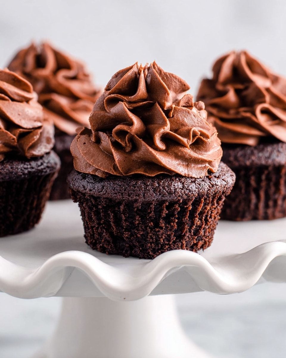 The image shows three chocolate cupcakes on a white cake stand with a wavy edge, set against a white marbled surface. Each cupcake has two visible layers: the base is a dark, moist chocolate cake with a slightly rough texture, and the top layer is a thick swirl of smooth, rich chocolate frosting with a light, fluffy texture forming soft ruffles. The cupcakes are closely placed, filling most of the frame, and the lighting highlights the chocolate's depth and creamy frosting. Photo taken with an iphone --ar 4:5 --v 7