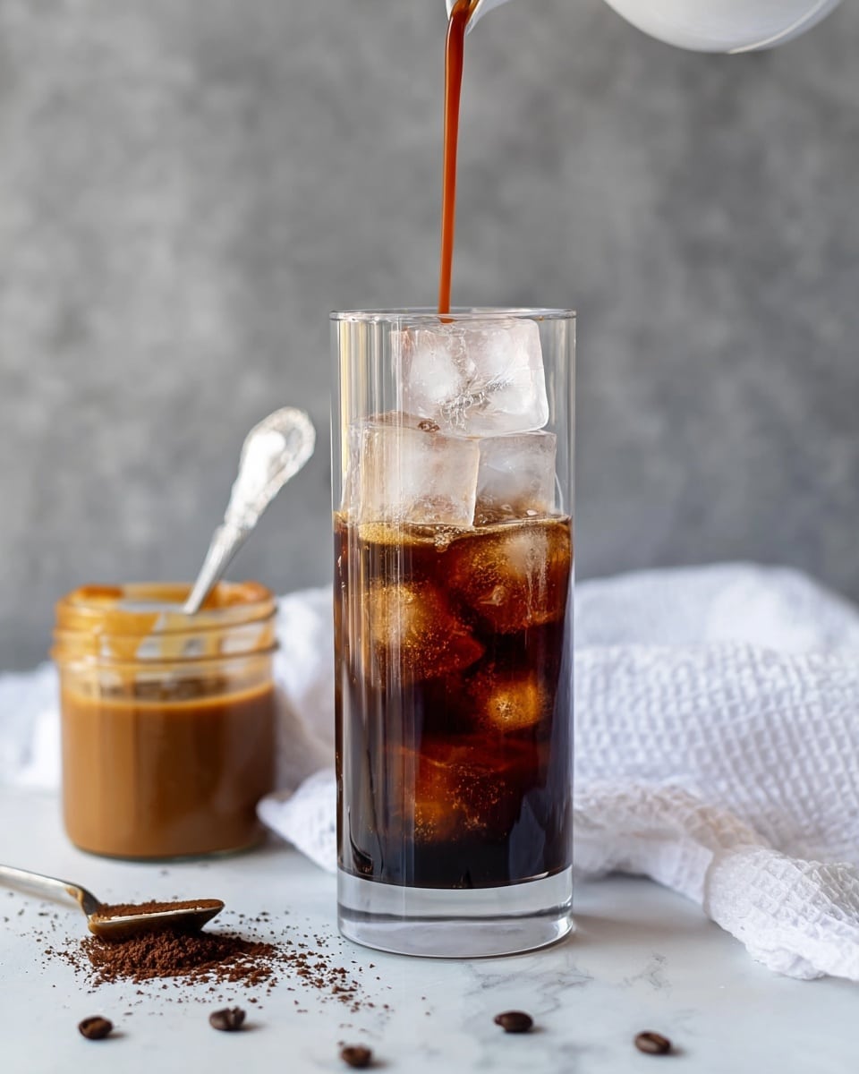 A tall clear glass filled halfway with large, slightly melting ice cubes, with dark brown coffee being poured into it from above, creating bubbles on the surface. To the left, a white jar contains caramel-colored sauce with a silver spoon inside it, resting on a white textured cloth. Coffee grounds are scattered lightly on the white marbled surface below the glass. The background is a soft gray with a subtle texture. Photo taken with an iphone --ar 4:5 --v 7