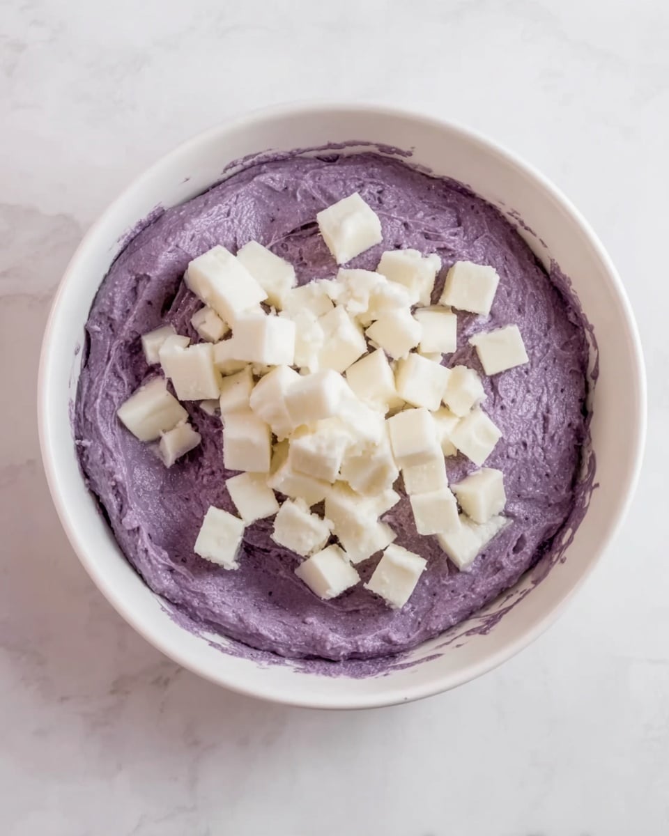 A white round bowl contains two layers of ingredients. The bottom layer is a thick purple mixture with a smooth but slightly rough texture, filling most of the bowl. On top of this purple base, there are many small white cubes scattered evenly, adding contrast and chunkiness. The bowl is placed on a white marbled surface. photo taken with an iphone --ar 4:5 --v 7