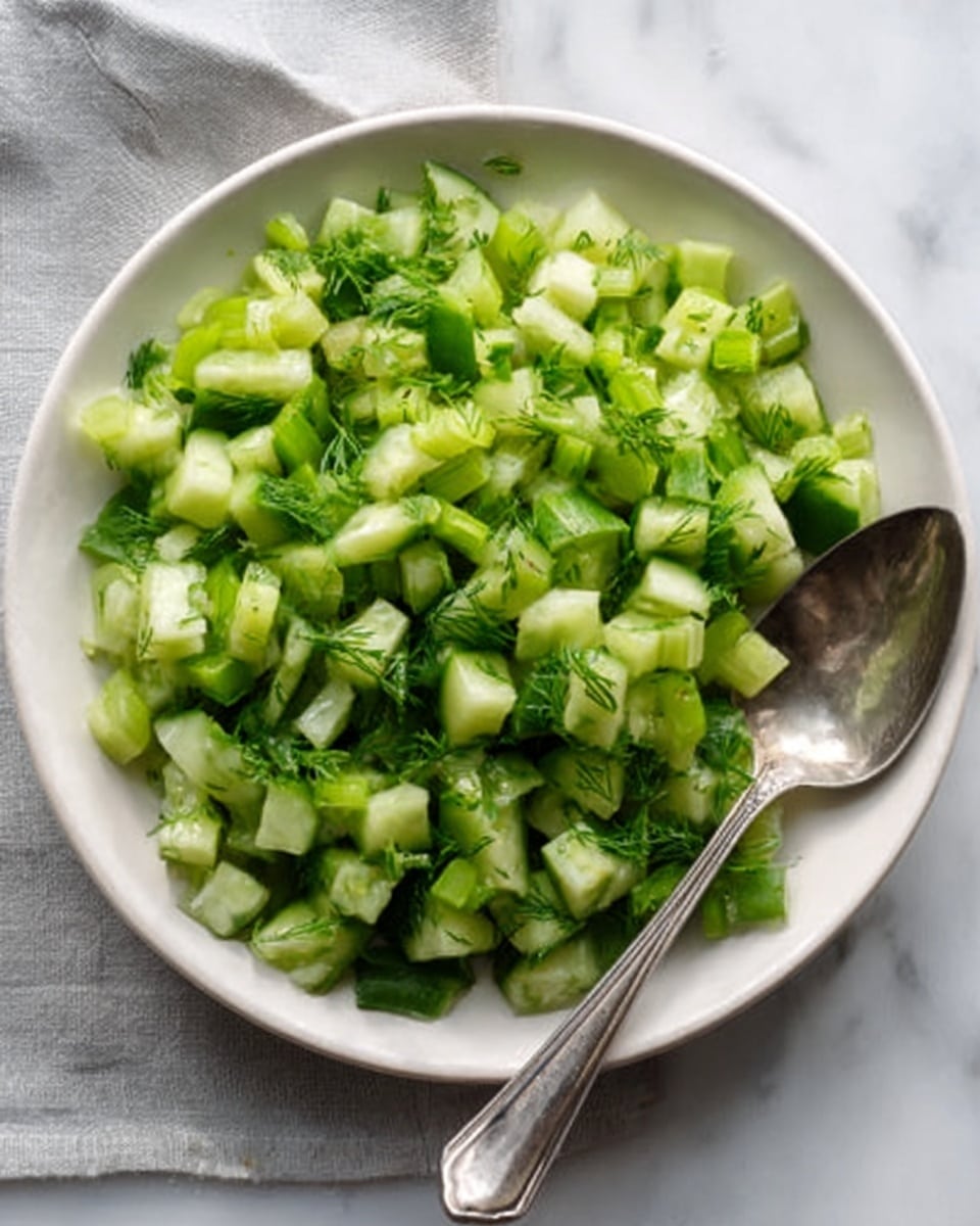 A white plate filled with a fresh salad made of small green cucumber pieces, chopped celery, and finely cut dill, all mixed together. The green vegetables vary in shades from light to dark green, creating a fresh and healthy look. A silver spoon with a shiny surface rests on the side of the plate. The plate is placed on a white marbled surface with a light gray cloth partially visible on the top left. Photo taken with an iphone --ar 4:5 --v 7