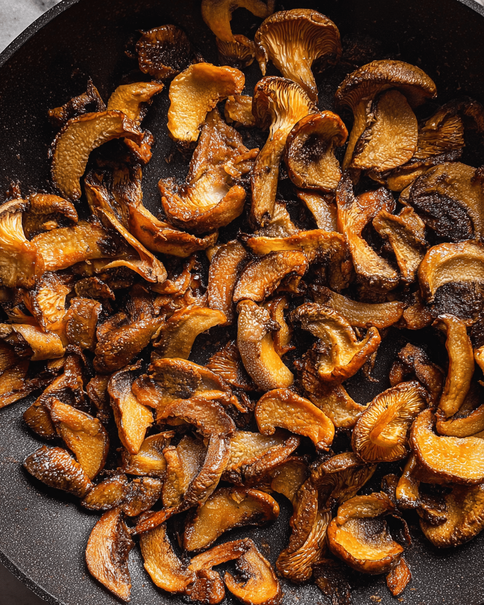 A close-up top view of a single layer of cooked mushroom slices in a black pan. The mushrooms are golden brown with darker caramelized spots, showing a mix of curved and flat shapes, some pieces with wrinkled texture and others smooth. The pan’s dark surface contrasts with the shining, slightly oily mushroom edges and the white marbled surface underneath is partly visible around the pan edges. photo taken with an iphone --ar 4:5 --v 7
