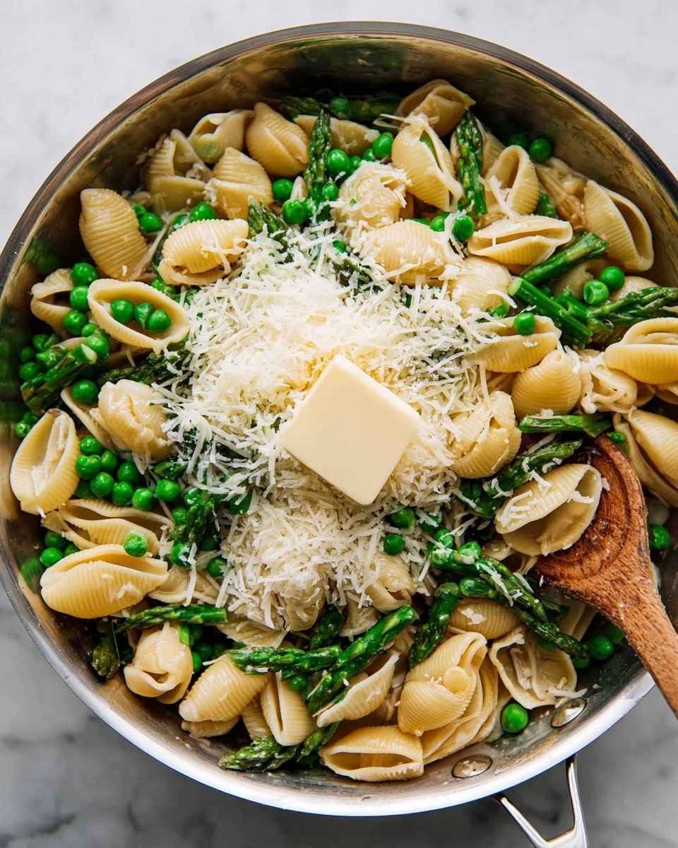 A metal pan filled with cooked shell pasta mixed with bright green peas and sliced green asparagus scattered throughout. On top of the pasta, there is a mound of shredded white cheese covering part of the pasta with a square of pale yellow butter placed in the center. A wooden spoon is resting on the right side of the pan, partially under the pasta and vegetables, with some pasta on it too. The pan is placed on a white marbled surface. photo taken with an iphone --ar 4:5 --v 7