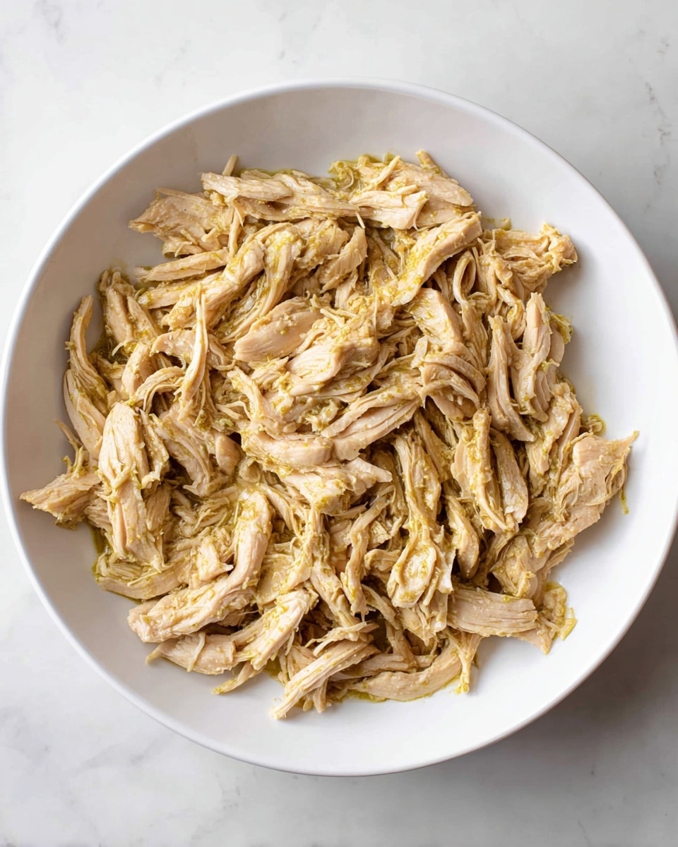 A white plate is filled with shredded cooked chicken pieces that are light beige with a slight yellow tint from a sauce or seasoning. The chicken is arranged loosely covering the whole surface of the plate, showing varied textures from smooth meat strips to some fibrous edges. The plate rests on a white marbled surface, adding a clean and simple background to the dish. photo taken with an iphone --ar 4:5 --v 7