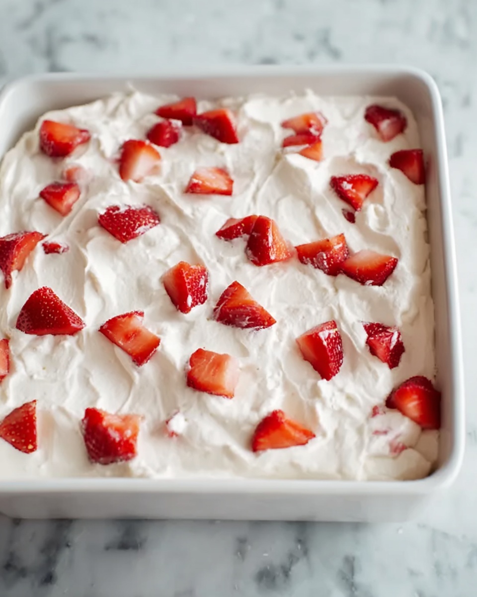 The image shows a square white dish filled with a dessert that has two main visible layers. The bottom layer is not shown clearly but is covered by a thick top layer of smooth, white whipped cream spread evenly across the dish. On top of the whipped cream, there are small pieces of fresh, bright red strawberries scattered randomly. The white marbled surface beneath the dish adds a clean and simple look. Photo taken with an iphone --ar 4:5 --v 7