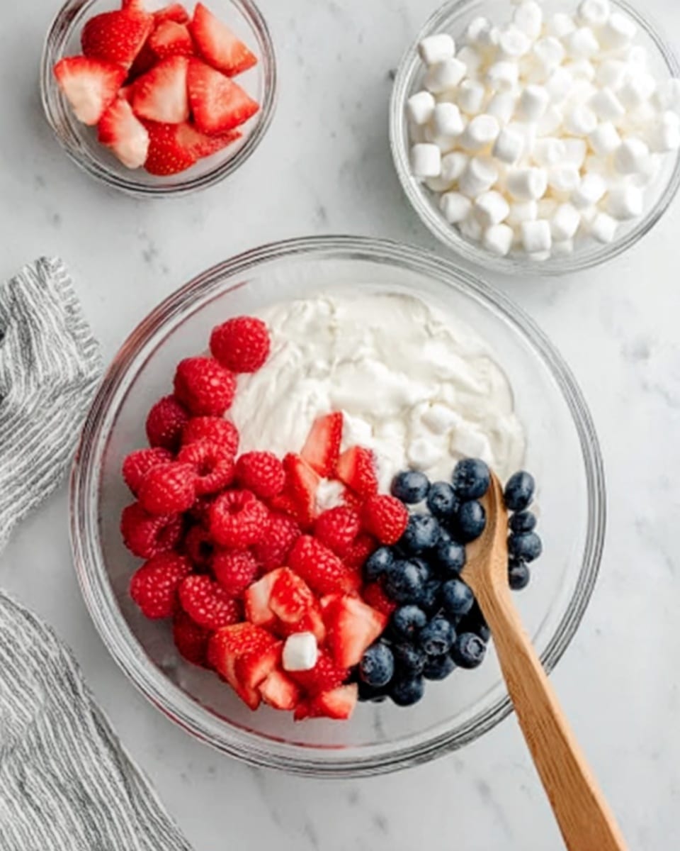 The image shows a clear glass bowl on a white marbled surface. Inside the bowl, there are four visible layers: white yogurt covering half of the bowl's bottom, fresh red raspberries placed in a cluster on one side, sliced strawberries in small pieces near the raspberries, and a small handful of dark blue blueberries scattered next to the raspberries. A wooden spoon rests inside the bowl on the right side, with a woman's hand holding it. Nearby, there are two small clear glass bowls, one filled with white mini marshmallows and the other with sliced strawberries. A white and gray striped towel lies under the bowl on the left. photo taken with an iphone --ar 4:5 --v 7