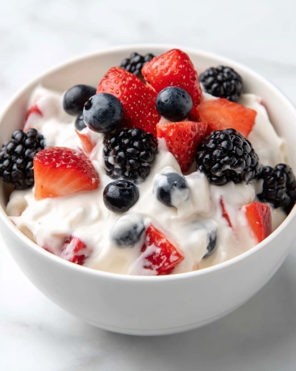 A white bowl filled with creamy yogurt mixed with pieces of strawberries, blackberries, and blueberries. On top, a small pile of fresh strawberries cut into chunks, shiny blackberries, and round blueberries forms a colorful layer. The yogurt looks smooth and thick, slightly covering some of the fruit pieces, while the white marbled surface beneath the bowl adds a clean, bright background. Photo taken with an iphone --ar 4:5 --v 7