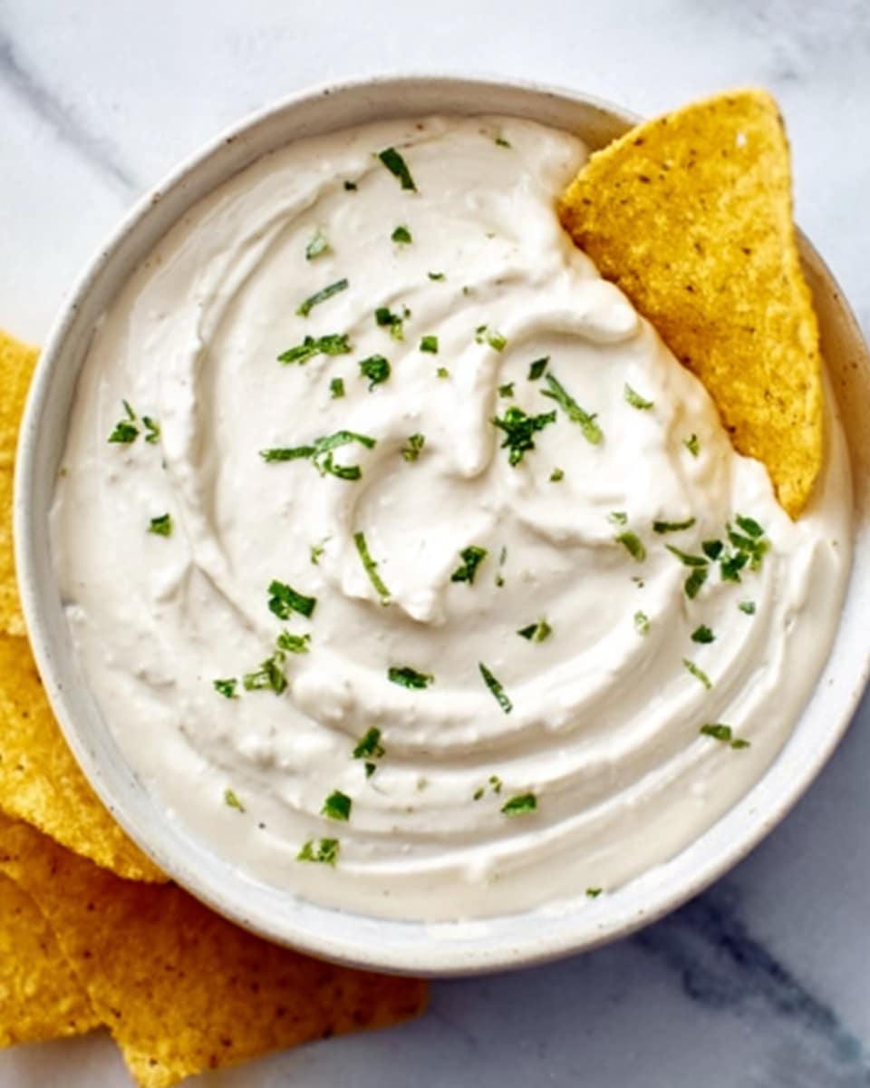 A white bowl filled with thick, creamy white dip with a smooth texture and slight swirls on top, garnished with small green herb pieces scattered evenly. A yellow tortilla chip is dipped into the side of the dip, resting on the bowl's edge. The bowl sits on a white marbled surface. photo taken with an iphone --ar 4:5 --v 7