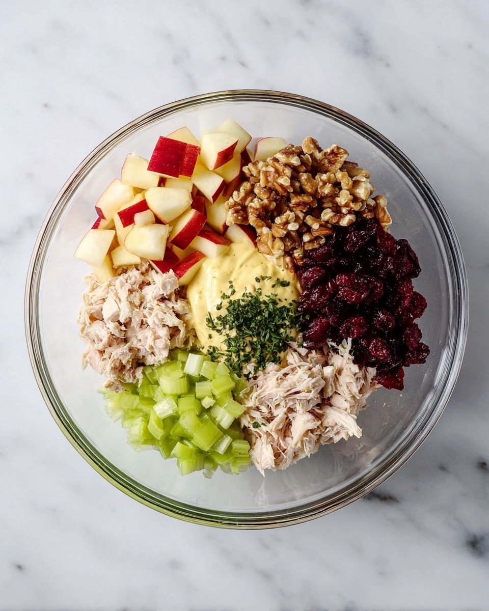 The image shows a clear glass bowl on a white marbled surface with six distinct sections of ingredients arranged neatly inside. Starting from the top left, there are small red and yellow apple cubes with a crisp texture, moving clockwise to chopped walnuts with a rough, crunchy look. Next is a smooth, pale yellow mayonnaise or dressing forming a semi-thick layer, followed by dark red dried cranberries with a wrinkled texture. Then, there is light pink, shredded chicken in soft chunks, and finally bright green diced celery with a fresh, crisp appearance. In the middle of the bowl, some finely chopped dark green herbs are placed. Photo taken with an iphone --ar 4:5 --v 7