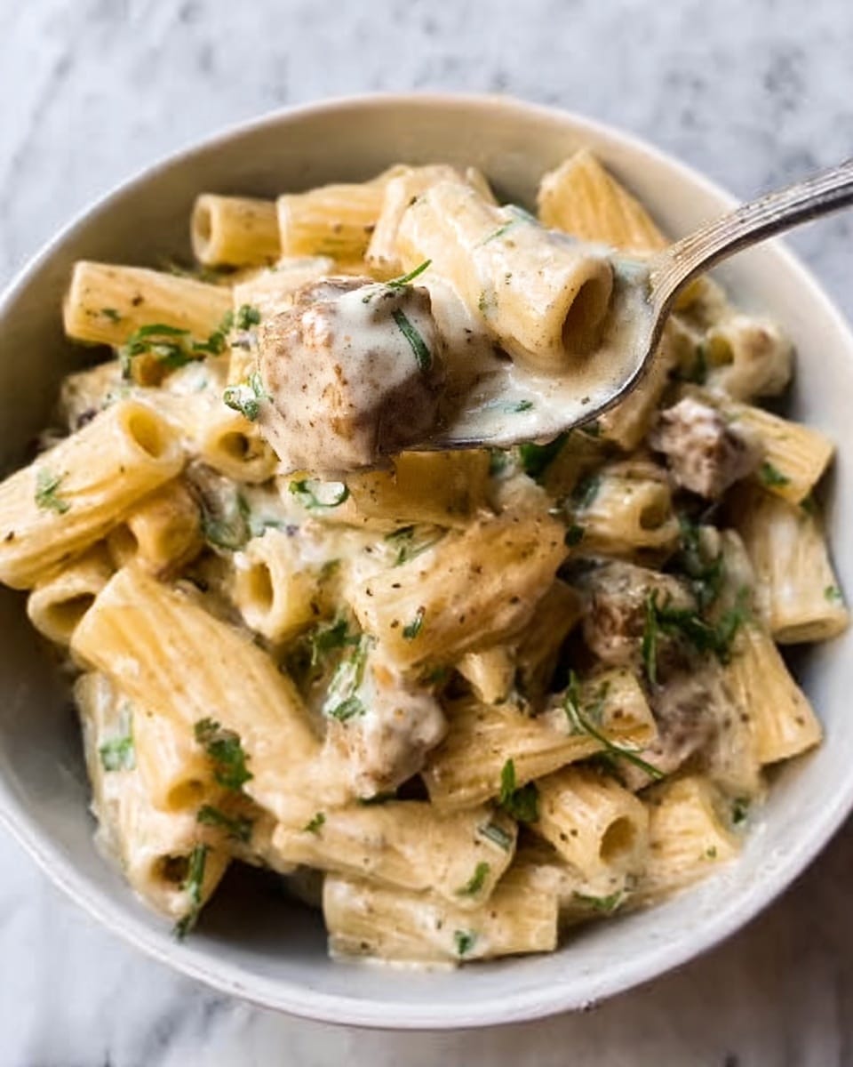 A white bowl filled with short tube pasta covered in a creamy white sauce with small green herb pieces scattered throughout, and chunks of light brown meat mixed in. A spoon is lifting some pasta from the bowl, showing the thick sauce and herbs clinging to the pasta. The bowl is set on a white marbled surface photo taken with an iphone --ar 4:5 --v 7