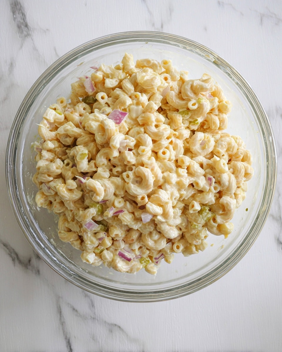 A clear glass bowl filled with creamy macaroni salad, showing about two layers of small elbow macaroni mixed with chopped boiled eggs and tiny bits of red onion all coated in a pale yellow creamy dressing with some green pickle pieces visible, placed on a white marbled surface. photo taken with an iphone --ar 4:5 --v 7