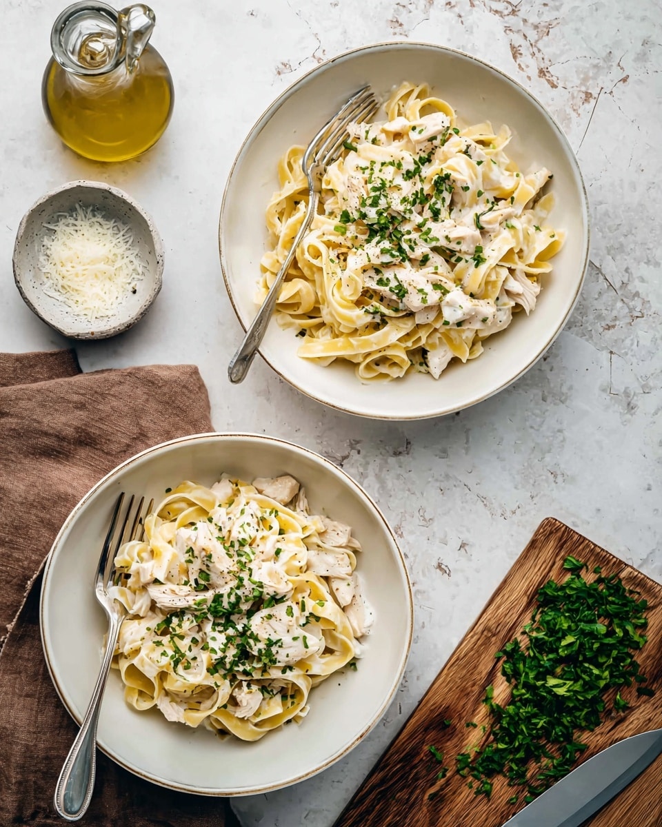 Two white bowls hold creamy pasta with three layers each: at the bottom creamy pale yellow fettuccine, in the middle white chunks of chicken, and on top finely chopped green herbs sprinkled evenly. Each bowl has a silver fork partly inside the pasta. To the left, a small bowl with white grated cheese and a glass bottle of olive oil sit on the white marbled texture surface. To the right, a wooden cutting board with chopped greens and a knife rests on the same surface, with a brown napkin nearby. Photo taken with an iphone --ar 4:5 --v 7