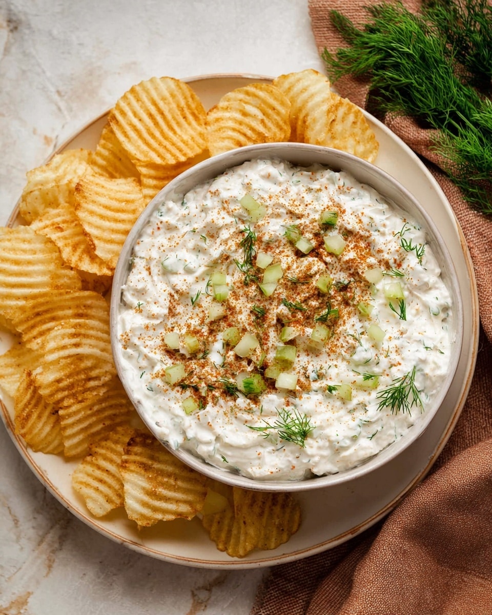 A white bowl filled with a creamy white dip that has a slightly thick texture, topped with small green dill sprigs, tiny light green pickle pieces, and a generous dusting of reddish-brown seasoning sprinkled evenly across the surface. The bowl sits on a white plate that holds a ring of ridged potato chips arranged around the bowl's edge. The whole setup is placed on a white marbled textured background with a soft brown cloth and fresh dill on the right side. Photo taken with an iphone --ar 4:5 --v 7