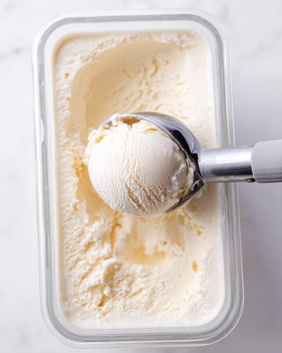 A close-up of a clear rectangular glass container filled with smooth, creamy vanilla ice cream. A metal ice cream scoop with a gray handle is lifting a round, perfectly shaped scoop from the center of the ice cream. The ice cream shows a soft, slightly textured surface with a pale ivory color. The background and surface are white marbled texture. photo taken with an iphone --ar 4:5 --v 7