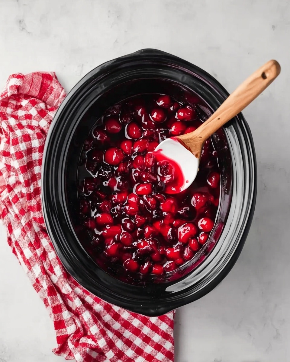 A black slow cooker filled with bright red, shiny cherry mixture, showing whole cherries and thick syrup. A wooden spoon with a white scoop is resting inside, coated with the cherry sauce. Next to the cooker is a folded red and white checkered cloth on a white marbled surface. Photo taken with an iphone --ar 4:5 --v 7