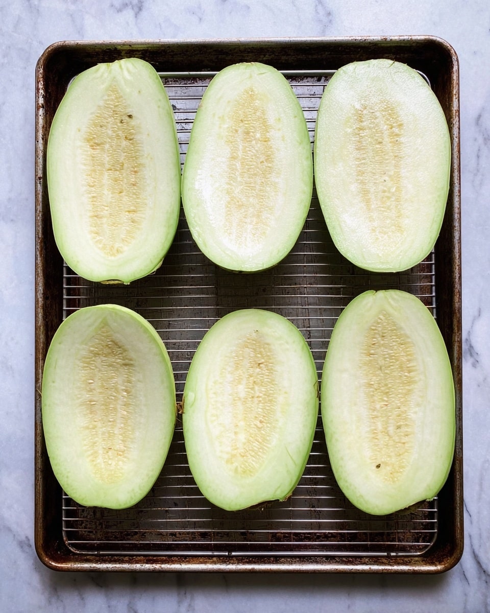 Six large slices of pale green eggplant are neatly placed on a metal rack inside a baking tray, arranged in two rows of three. Each slice is thick, showing smooth, light flesh with tiny seeds in the center and a slightly glossy texture. The baking tray has a worn, dark metal appearance and sits on a white marbled surface. photo taken with an iphone --ar 4:5 --v 7