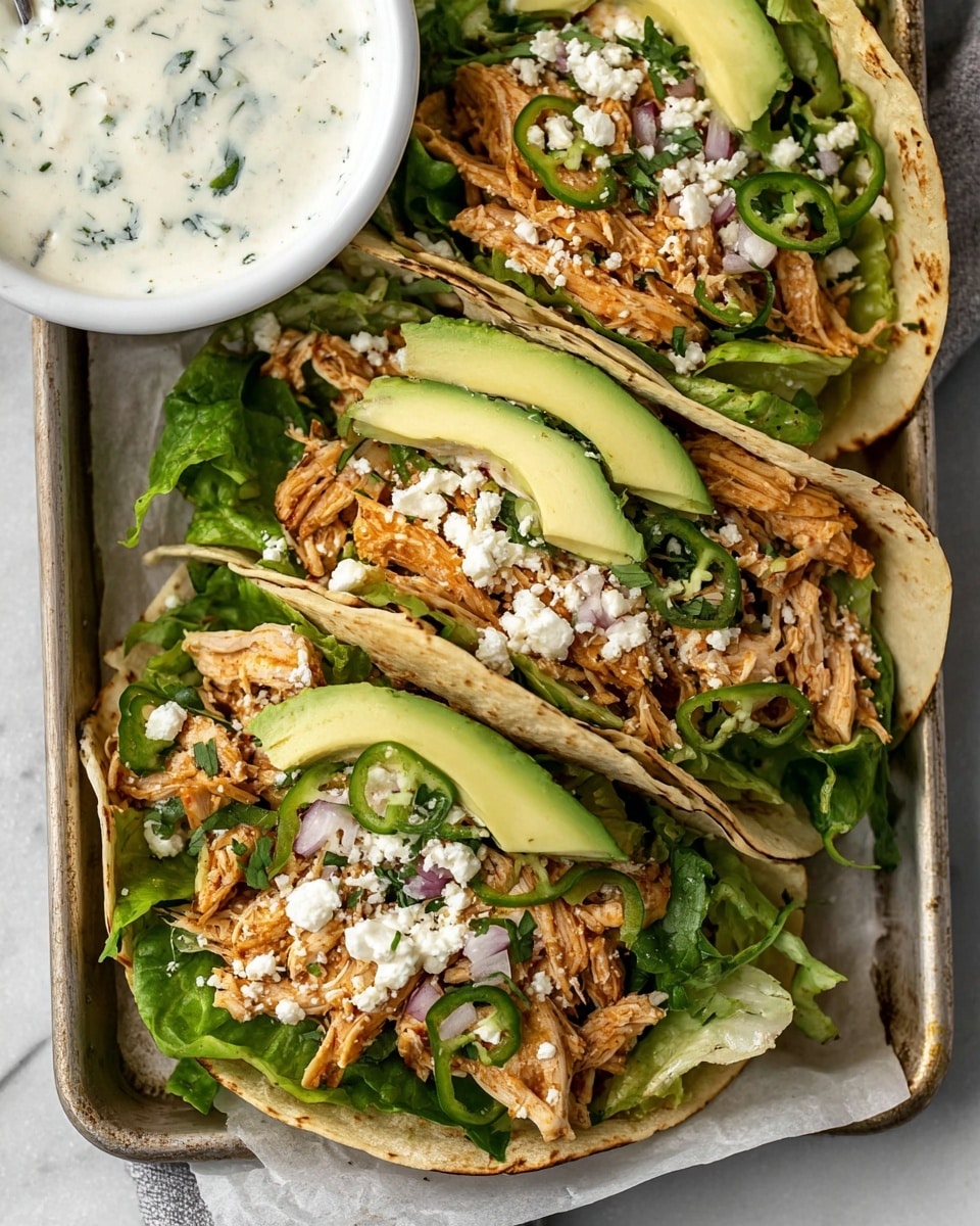 Three soft tortillas lie on a white marbled surface, each one filled with several layers. The base is filled with bright green lettuce leaves, topped with shredded light brown chicken mixed with small pieces of red onions and green chili slices. Thin slices of avocado with a creamy texture are placed on one side of each tortilla. Crumbled white cheese is sprinkled over the chicken and greens. At the top left corner, a white bowl holds a creamy white sauce with small green herb bits. The whole scene is set on a metal tray, enhancing the warm and fresh feel of the food. Photo taken with an iphone --ar 4:5 --v 7