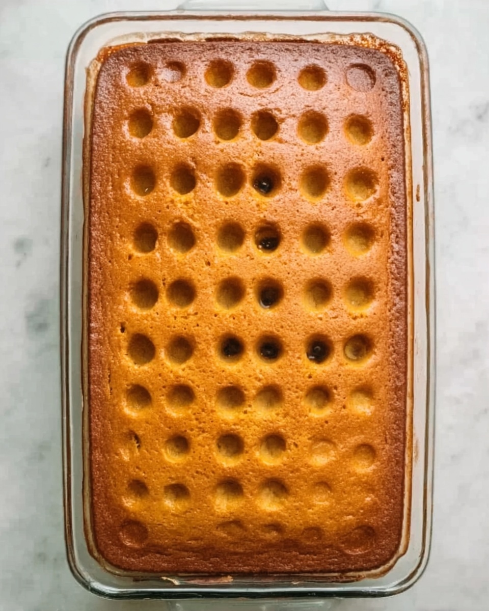 A rectangular glass baking dish with a golden brown cake inside, the cake has evenly spaced round holes all over the top, showing a moist texture. The edges of the cake are slightly darker and crispier, and the top surface is smooth with a slight shine. The dish sits on a white marbled surface. photo taken with an iphone --ar 4:5 --v 7