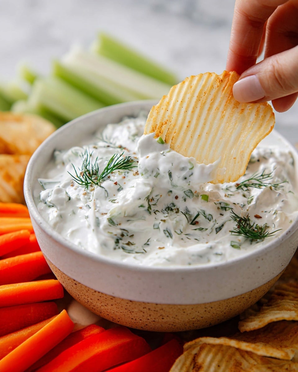 A woman's hand is holding a light-colored ridged potato chip dipped in thick, creamy white dip with green herbs mixed in, including small pieces of chives and sprigs of dill. The dip is in a round white bowl with a slightly rough texture on the outside. Around the bowl, there are bright orange carrot sticks, red pepper pieces, and pale celery sticks, all laid out on a white marbled surface. The focus is on the chip dipping into the herb-filled creamy white dip. photo taken with an iphone --ar 4:5 --v 7