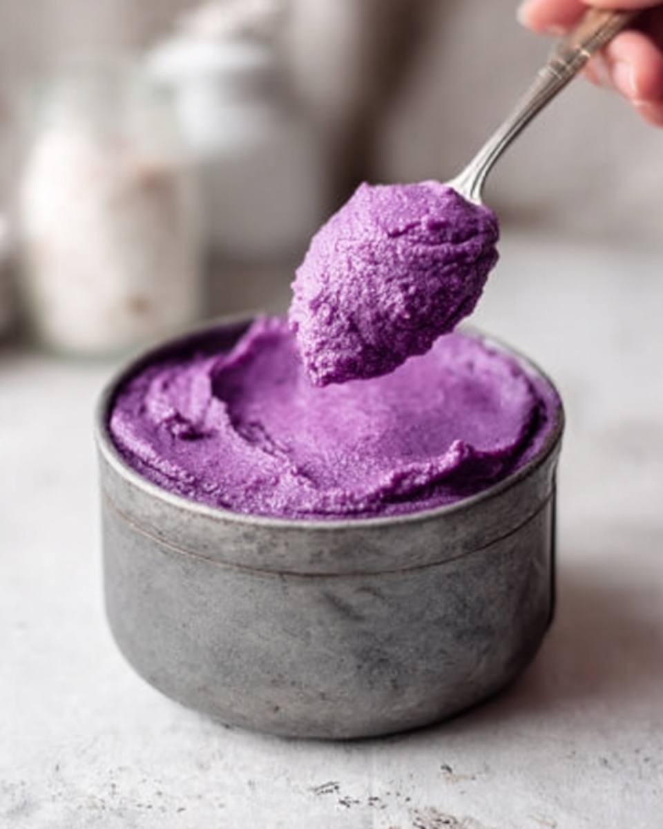 This image shows a single gray metal bowl filled with smooth purple mashed food, looking soft and creamy in texture. A silver spoon is scooping up a portion of the purple mash, with the spoon held by a woman's hand that is partly visible at the top left. The background and surface are a white marbled texture, with other blurred white items in the back, giving a clean kitchen feel. photo taken with an iphone --ar 4:5 --v 7